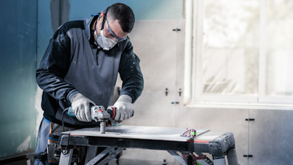 En person slipar ett metallstycke på ett arbetsbord med en eldriven slipmaskin, iklädd skyddsglasögon och handskar.