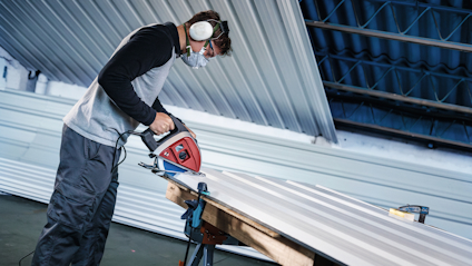 Person sågar i metallplåt med röd cirkelsåg, bär skyddsglasögon och hörselskydd, inomhusmiljö.