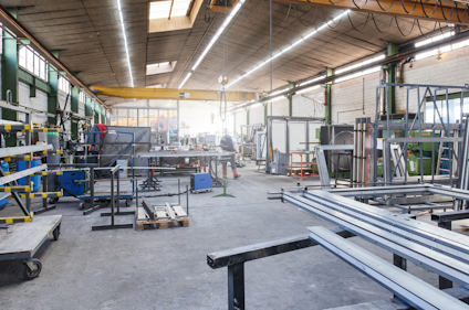 Verkstad med metallstänger, verktyg och industriutrustning. Takbelysning och stora fönster syns i bakgrunden.