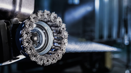 Roterande borste med flätade metalltrådar på en maskin, suddig bakgrund av metallisk yta.