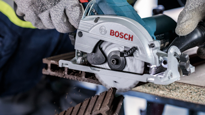 Cirkelsåg från Bosch sågar genom träskiva; händer i skyddshandskar syns.