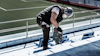 Person använder en borrmaskin på en stadionläktare klädd i hjälm och skyddsutrustning. Fotbollsplan i bakgrunden.