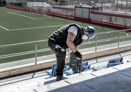 Person i arbete med borrmaskin på en läktare vid en fotbollsplan.