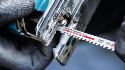 Jigsaw with Bosch "Expert Hardwood fast" blade, close-up of gloved hands holding tool.