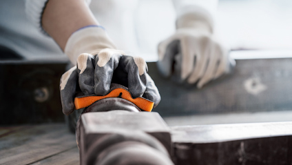 Händer i arbetshandskar som håller i metallverktyg på träyta.