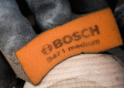 Arbetsvante med orange läderdel och texten "BOSCH ser i medium", handen håller något träaktigt.