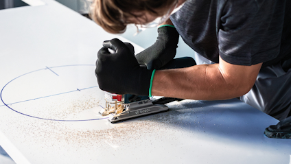 Person sågar cirkel i vitt material med sticksåg, iklädd svarta handskar.