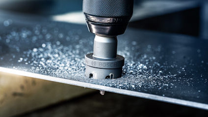 Metallborr med hålsåg borras genom en metallplåt, metallspån runt borren.