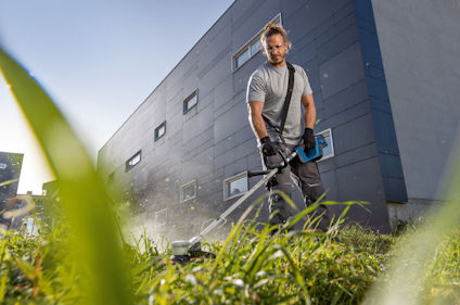 En man använder en bensintrimmer för att klippa gräs framför en modern byggnad. Gräs i förgrunden.