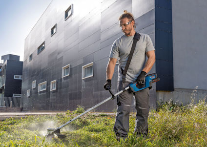 Man använder trimmer för att klippa gräs framför moderna byggnader.