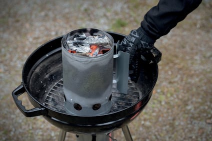 Engångsgrill med tändrör fylld med kol, tänd ovanpå en rund grill. Handske håller i tändröret.