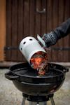 Kol hälls från en tändrörsstarter ner i en klotgrill av en person med grillvante.