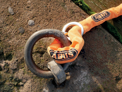 Orange kedjelås med Abus-logga, fastsatt runt en rostig järnring på en grov stenyta.