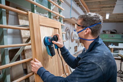 Person slipar trädörr med elektrisk slipmaskin inomhus. Bär skyddsglasögon och ansiktsmask.